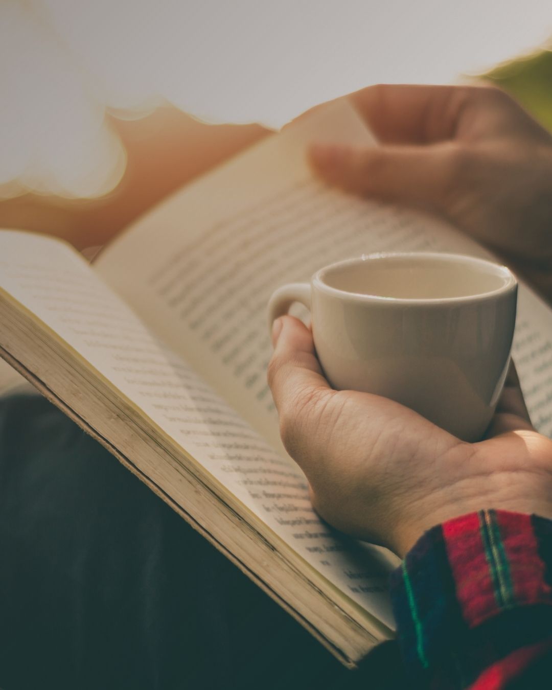 Handen houden een kop thee vast boven een opengeslagen boek in het zonlicht